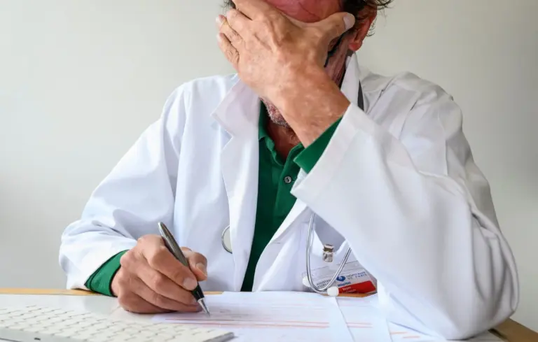 1444x920 an exhausted doctor in a white coat covers his face with his hand while filling out adminis